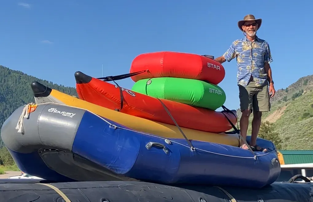 Man with cowboy hat standing on an inflatable raft loaded with colorful kayaks on the Snake River